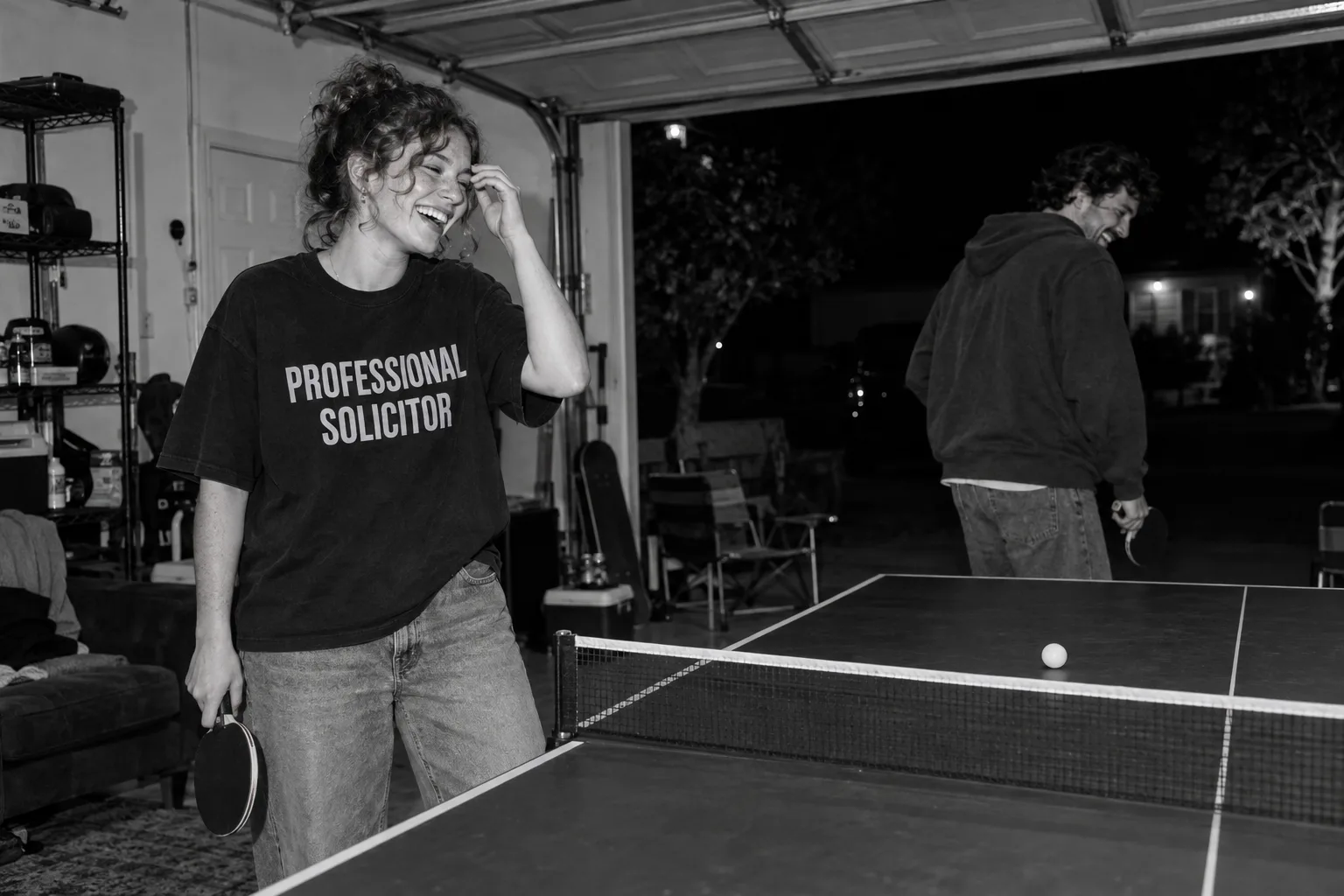 Candid campaign photograph of teammates laughing during table tennis; one wears an oversized Professional Solicitor tee.