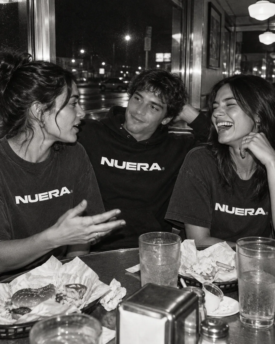 Three friends sharing a meal in a diner in NUERA tees and a hoodie.