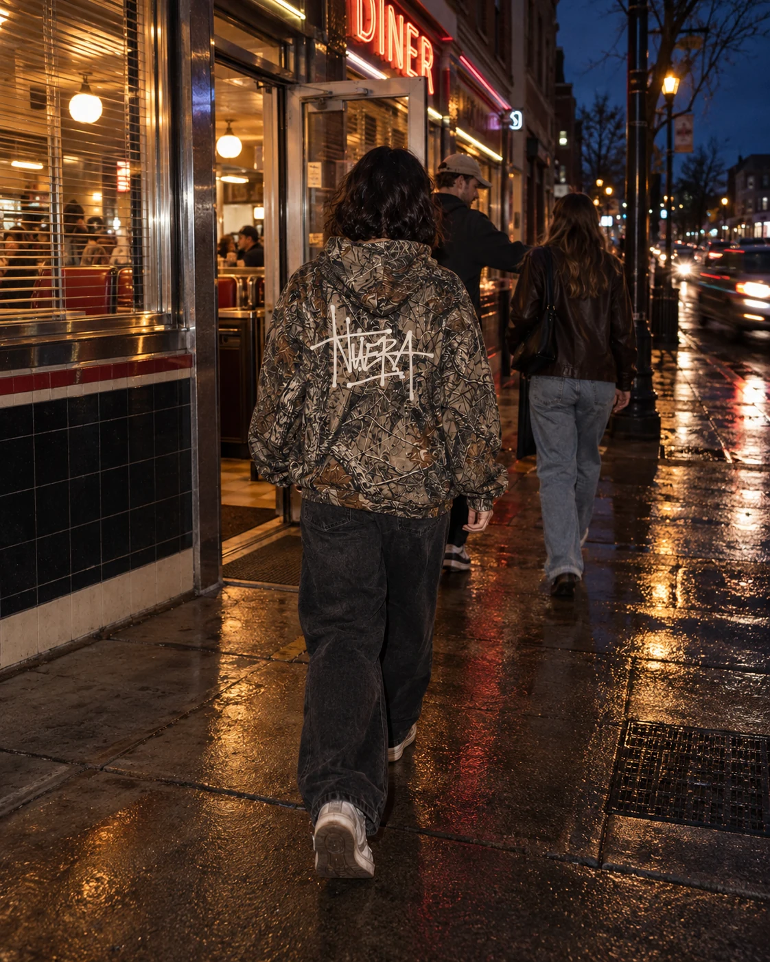 A person in a camouflage NUERA script hoodie following friends toward a diner at night.