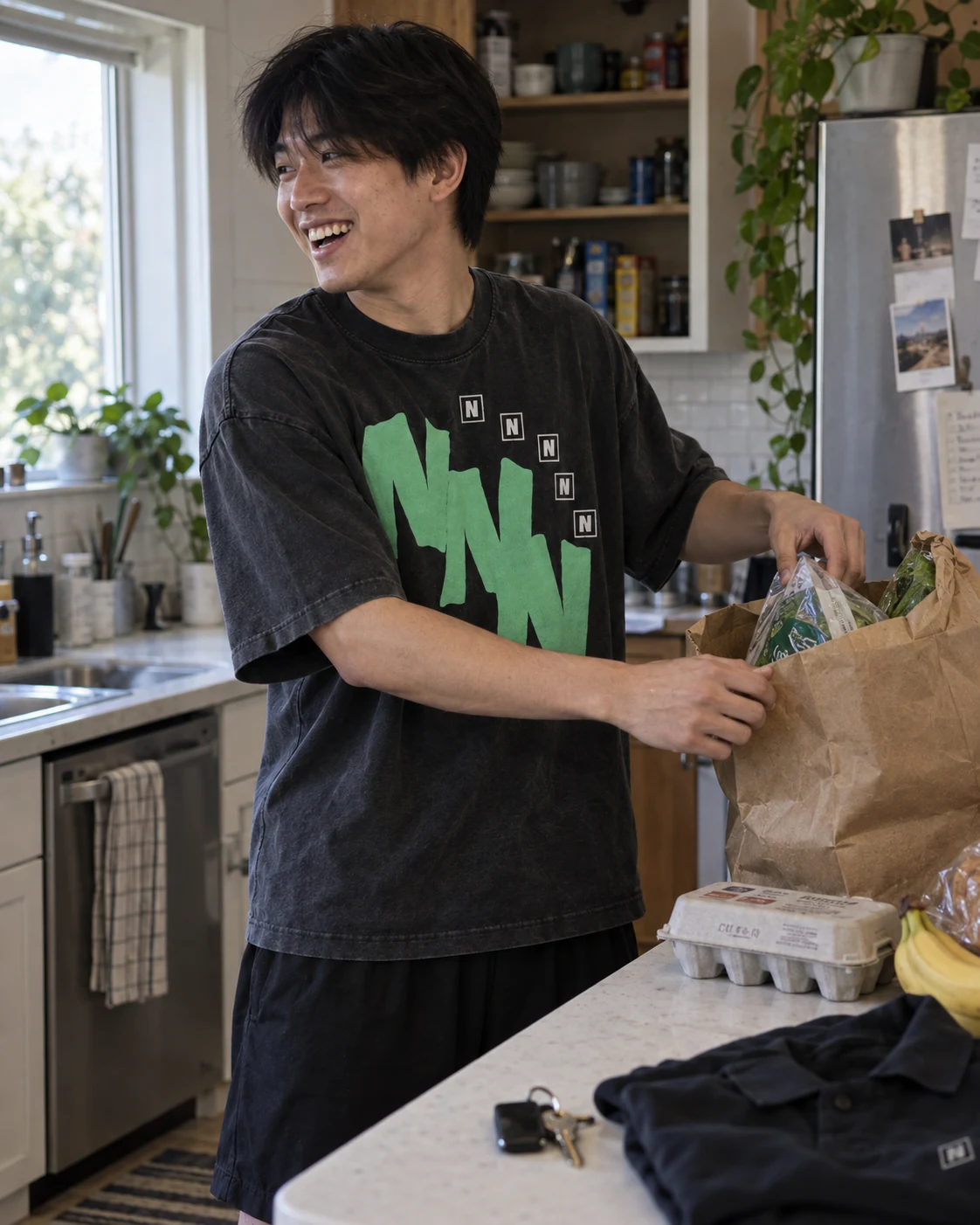 A person in an oversized mint NUERA graphic tee unpacking groceries at the kitchen counter.
