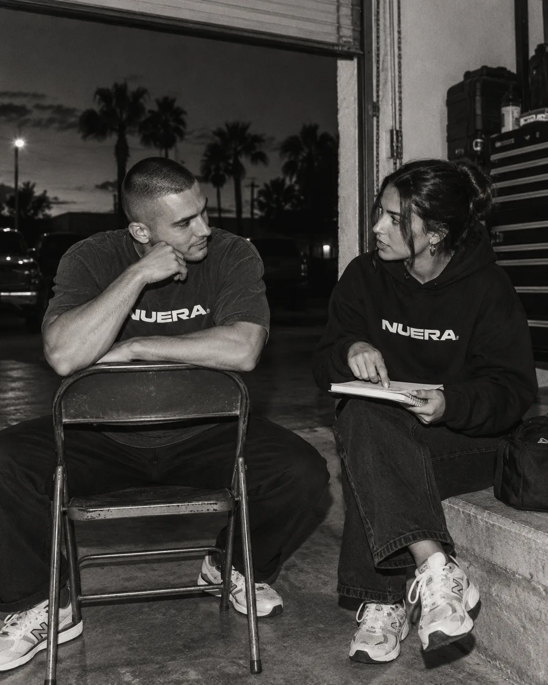 Two adults in a NUERA tee and hoodie discussing notes by an open garage door.